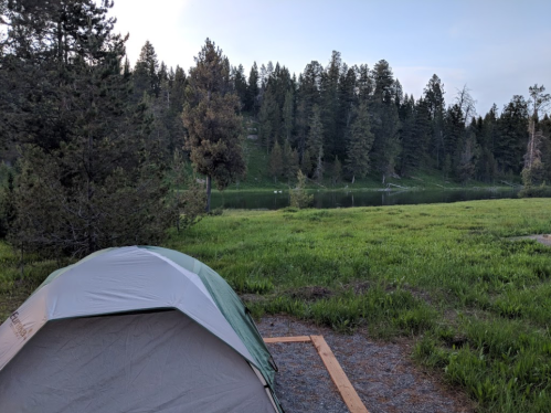 A tent set up near a serene lake, surrounded by lush green grass and tall trees under a clear sky.