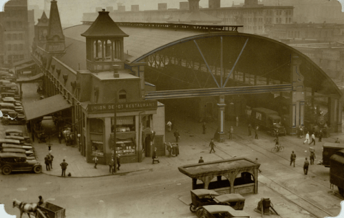 Historic black-and-white photo of a bustling train station with cars, people, and a restaurant building.