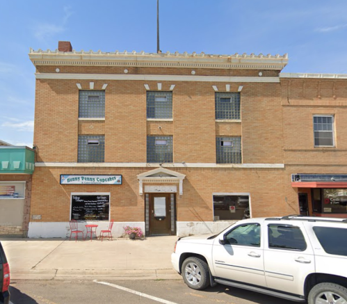 A brick building with three windows and a sign for "Penny Penny Cupcakes" on the ground floor, parked cars in front.