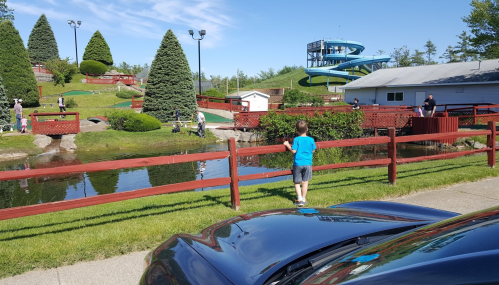 A child stands by a wooden fence, overlooking a pond with mini-golf and a water slide in the background on a sunny day.
