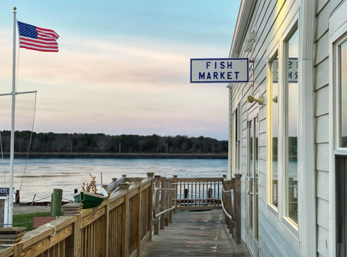 A wooden walkway leads to a fish market by the water, with an American flag waving in the breeze at sunset.