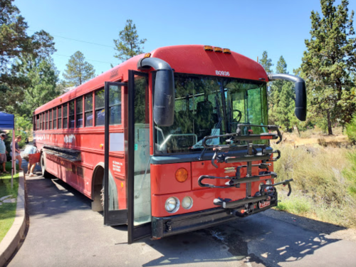 A red bus parked in a wooded area, featuring bike racks on the front and people in the background.