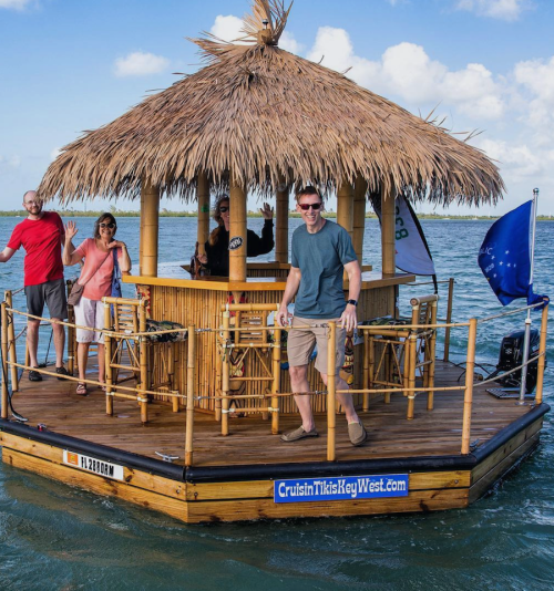 A group of people on a tiki boat with a thatched roof, enjoying the water and waving at the camera.
