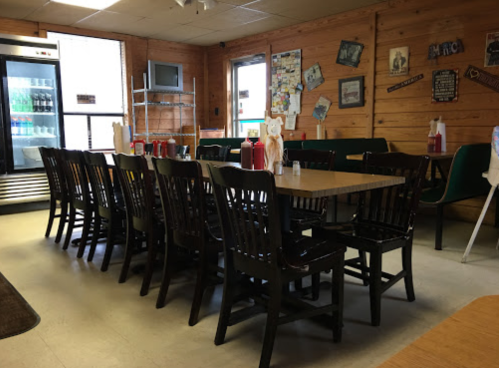 A rustic diner interior with wooden tables and chairs, ketchup bottles on tables, and a soda fridge in the background.