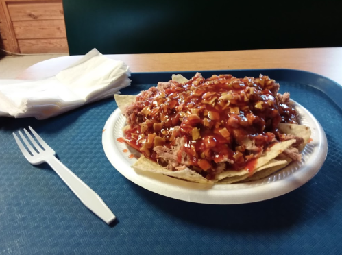 A plate of nachos topped with pulled pork, barbecue sauce, and diced toppings, served on a disposable tray.