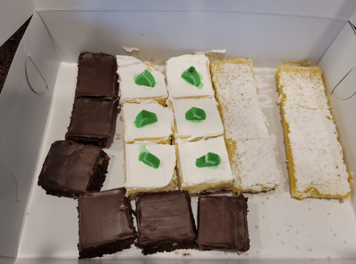 A box of assorted desserts, including chocolate squares, cream-topped squares with green jelly, and lemon bars.