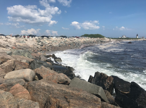 Rocky shoreline with waves crashing, cars parked nearby, and a lighthouse in the distance under a blue sky.
