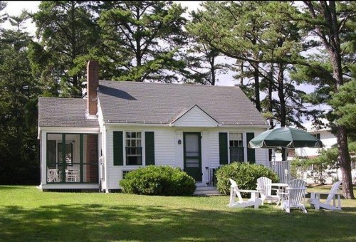 A charming white cottage with green shutters, surrounded by trees and featuring a patio with chairs and an umbrella.