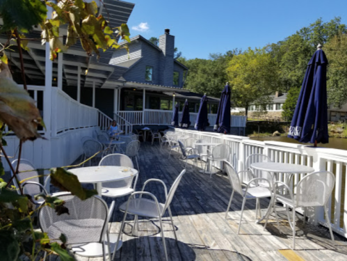Outdoor dining area with white tables and chairs, shaded by blue umbrellas, beside a calm water view.
