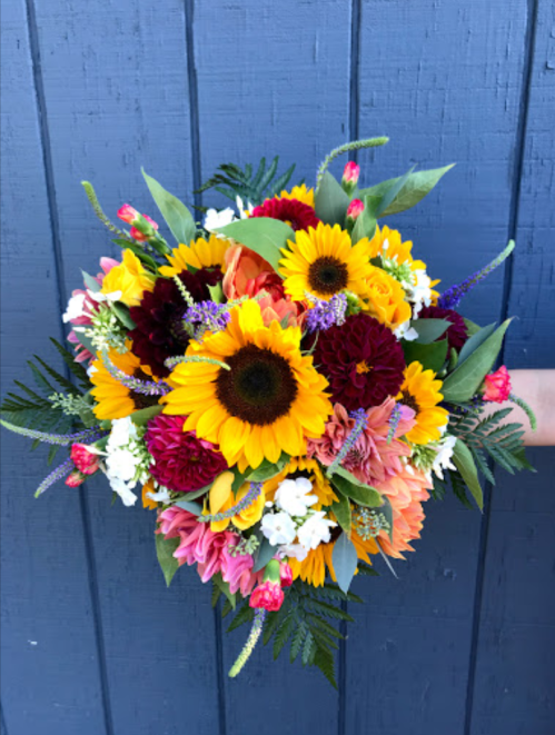 A vibrant bouquet featuring sunflowers, dahlias, and various colorful flowers against a blue wooden background.