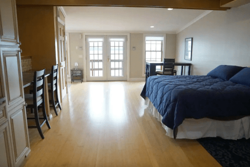 A spacious studio with a bed, dining table, and large windows, featuring wooden floors and a cozy atmosphere.