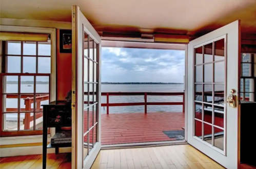 A view from an open doorway overlooking a calm body of water and a wooden deck, with cloudy skies in the background.