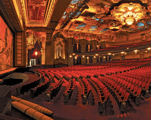 A grand theater interior with ornate decor, featuring rows of red seats and a large stage with a colorful backdrop.