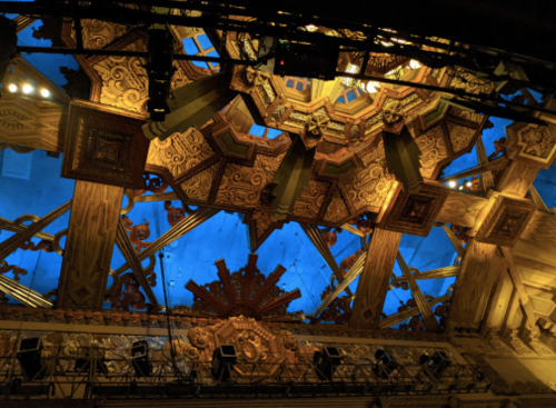 Intricate wooden ceiling with ornate designs and blue lighting, viewed from below.