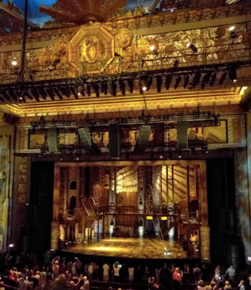 A grand theater stage illuminated with warm lighting, featuring ornate decorations and an audience gathering below.