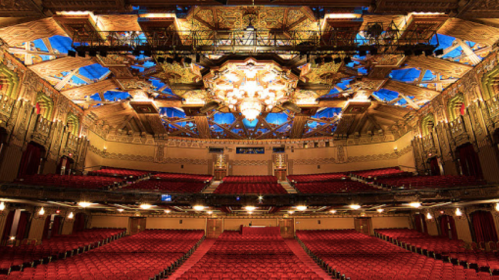 A grand theater interior with ornate architecture, a stunning chandelier, and rows of red seats under a colorful ceiling.