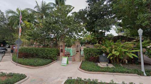 Entrance to a lush garden with palm trees, greenery, and a sign, set in a residential area.
