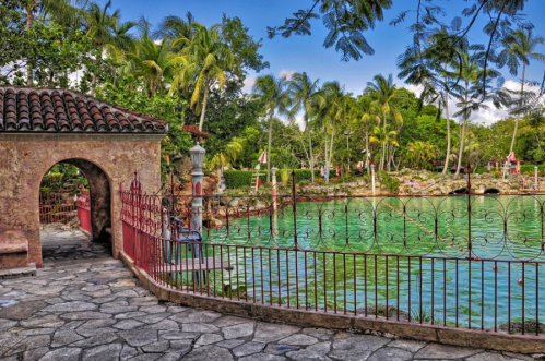 A scenic view of a lush garden with palm trees, a turquoise pond, and a stone pathway leading to an archway.