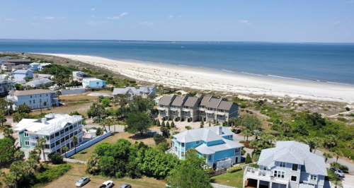 Aerial view of coastal homes near a sandy beach and calm blue water under a clear sky.