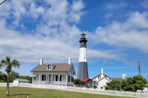 A black and white lighthouse beside a white house, surrounded by greenery and a blue sky with fluffy clouds.