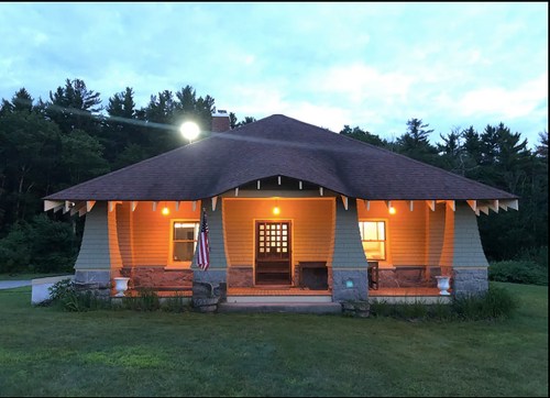 A cozy, illuminated house with a unique roof design, surrounded by trees at dusk, featuring an American flag.