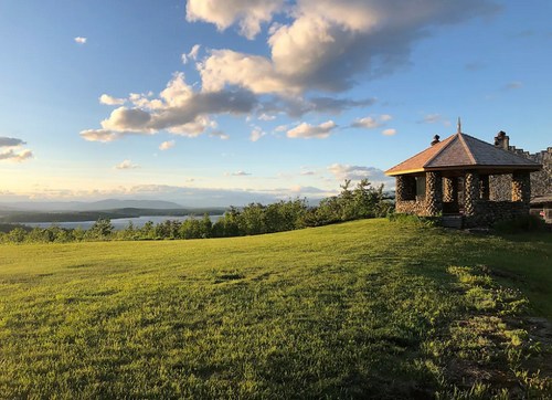 A scenic view of a stone gazebo on a grassy hill, overlooking a lake and distant mountains under a blue sky with clouds.
