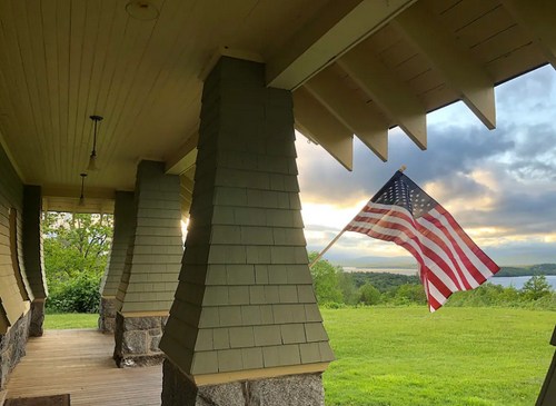 A porch with stone columns and a waving American flag, overlooking a green landscape and a lake under a cloudy sky.