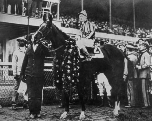 A historic black-and-white photo of a jockey on a horse adorned with a floral wreath, surrounded by a cheering crowd.