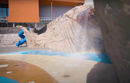 A water slide next to a rocky structure with a small waterfall, set in a recreational area.