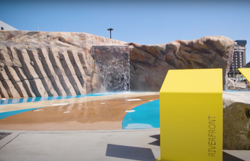A colorful water feature with a waterfall and rock formations, surrounded by a paved area and yellow signage.
