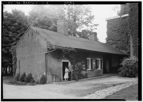 A historic brick building covered in ivy, with two people standing at the entrance on a gravel path.