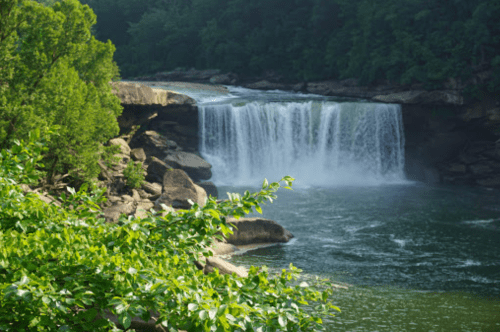 A serene waterfall cascades into a river, surrounded by lush greenery and rocky cliffs.