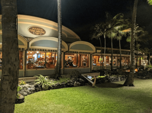 A restaurant with large windows, palm trees, and outdoor seating, illuminated at night.