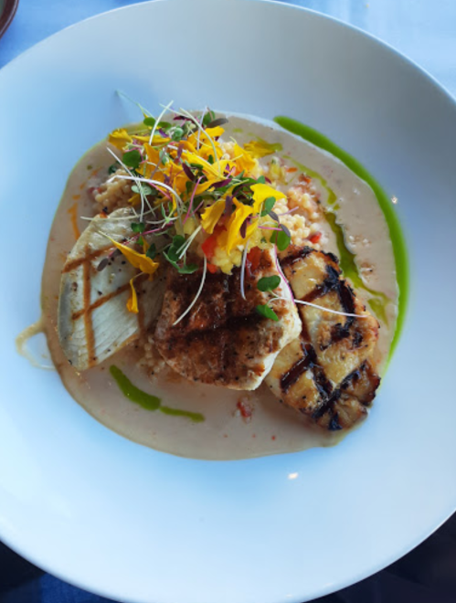 A beautifully plated dish featuring grilled fish, creamy risotto, and vibrant garnishes on a white bowl.