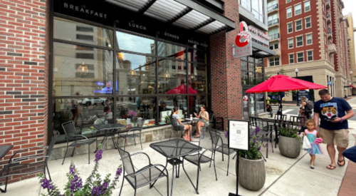 Outdoor dining area of a restaurant with tables, umbrellas, and people enjoying meals in a city setting.