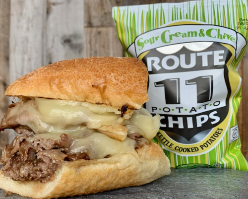 A hearty sandwich with melted cheese and roast beef beside a bag of Route 11 sour cream and chive potato chips.