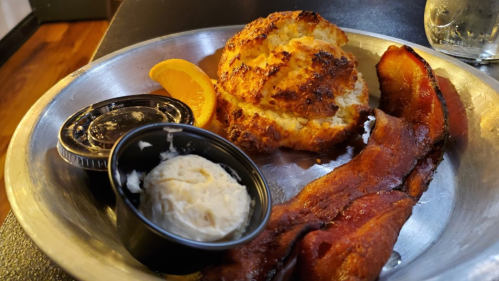 A metal plate with crispy bacon, a biscuit, butter, and an orange slice.