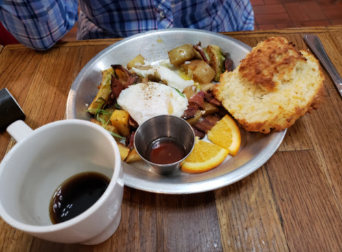 A plate of breakfast with potatoes, bacon, a poached egg, orange slices, and a biscuit, alongside a cup of coffee.