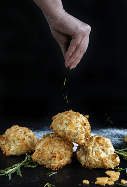 A hand sprinkles herbs over a stack of golden, flaky biscuits on a dark background, with fresh rosemary scattered around.
