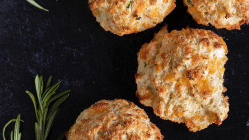 Four golden-brown biscuits with a flaky texture, garnished with fresh rosemary on a dark surface.