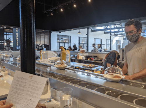 A man in a mask serves food at a modern restaurant with a counter and patrons in the background.