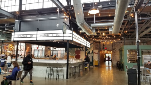 Interior of a modern food hall with a welcoming sign, communal seating, and industrial decor. Warm lighting enhances the atmosphere.