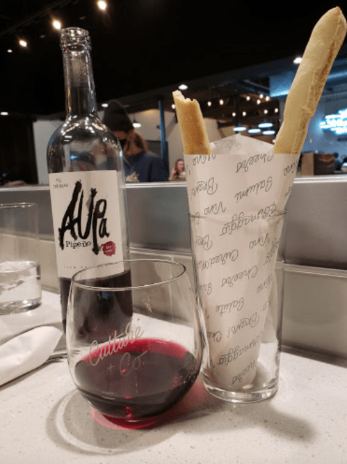 A bottle of red wine next to a glass of wine and a cup of breadsticks wrapped in paper at a restaurant.