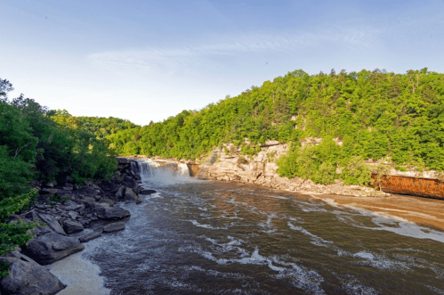 A serene waterfall cascades into a river, surrounded by lush green trees and rocky banks under a clear blue sky.