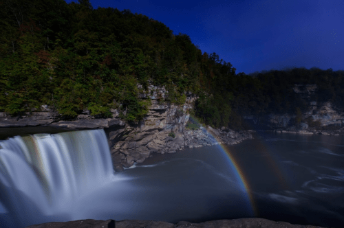 A serene waterfall cascades into a river, surrounded by lush greenery and a faint rainbow in the mist at night.