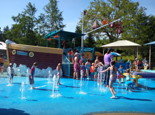 Children play in a colorful splash pad with water features, surrounded by families and a pirate-themed play structure.
