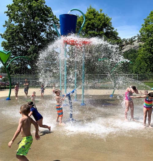 Children play in a splash pad, enjoying water sprays and sunny weather at a park.