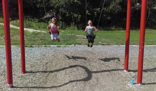 Two children joyfully swinging on red swings at a playground, surrounded by trees and a gravel surface.