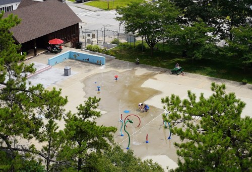 A splash pad with colorful water features, surrounded by trees and a building, with people enjoying the area.