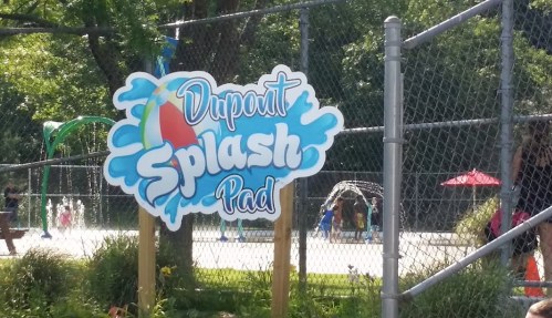 Sign for "Dropout Splash Pad" with water features and people enjoying the park in the background.